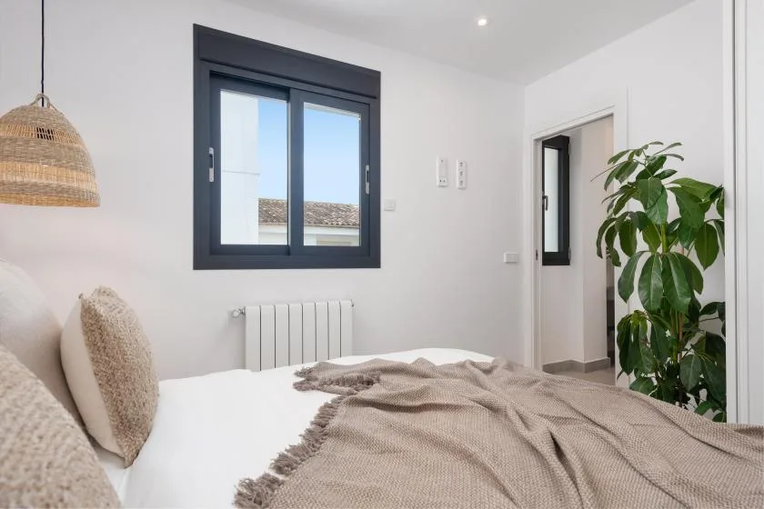 Un dormitorio luminoso con una cama, una ventana de marco oscuro y una planta frondosa.
