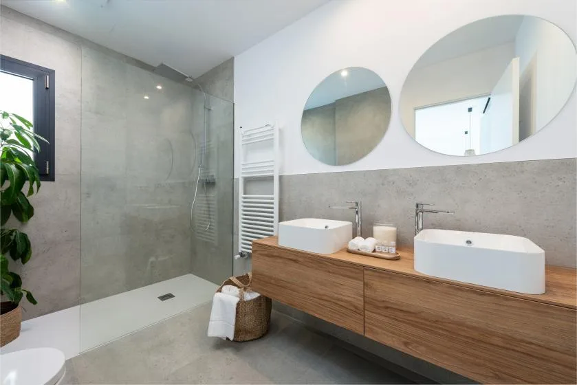 Un baño luminoso con ducha, lavabos dobles y espejos redondos.