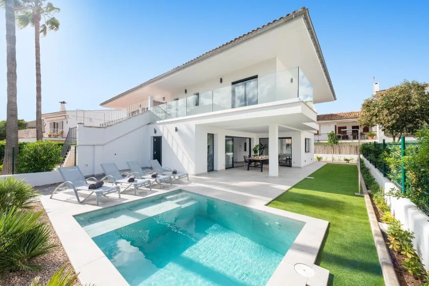 A modern white house overlooks a swimming pool, patio seating, and a neat lawn.