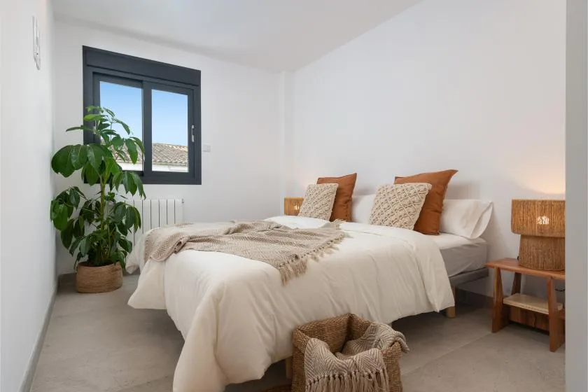 Un dormitorio luminoso con una cama bien hecha y una planta frondosa junto a la ventana.