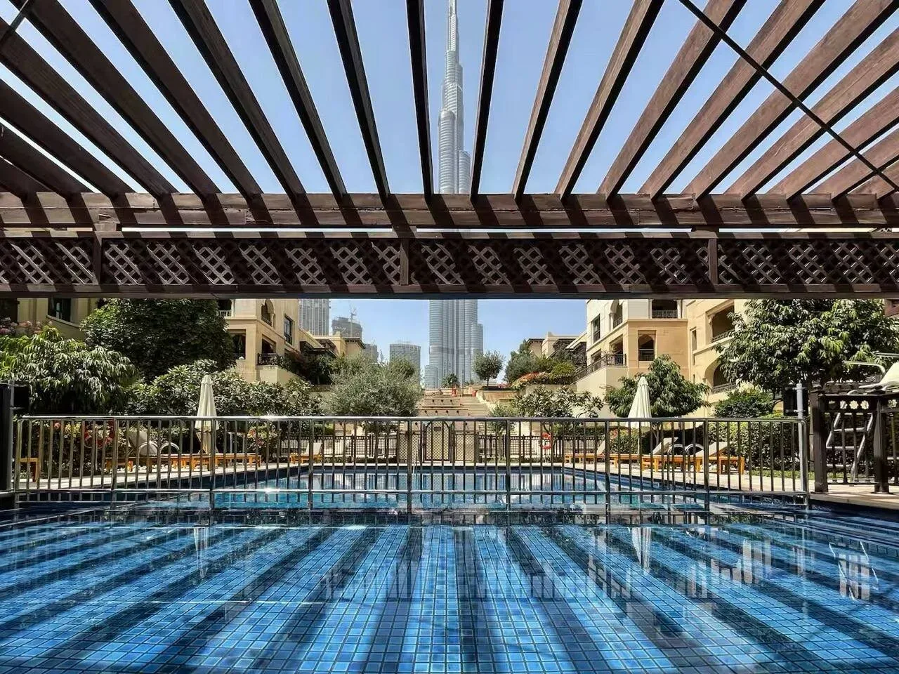 Una piscina de azulejos azules bajo una pérgola de madera, con edificios al fondo.