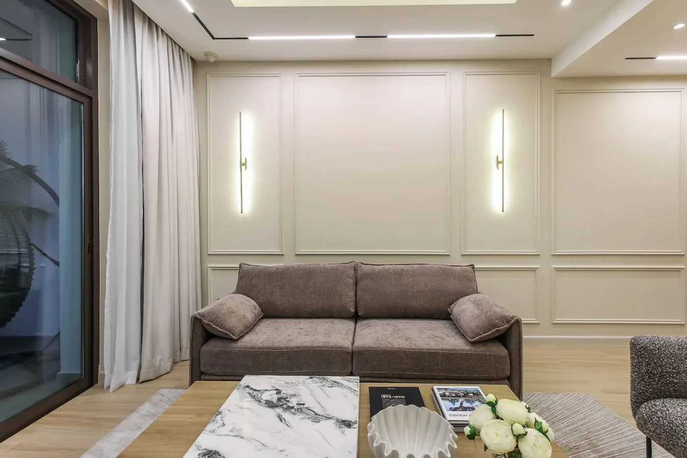 Una sala luminosa con un sofá gris, cortinas largas y una mesa de centro de mármol.