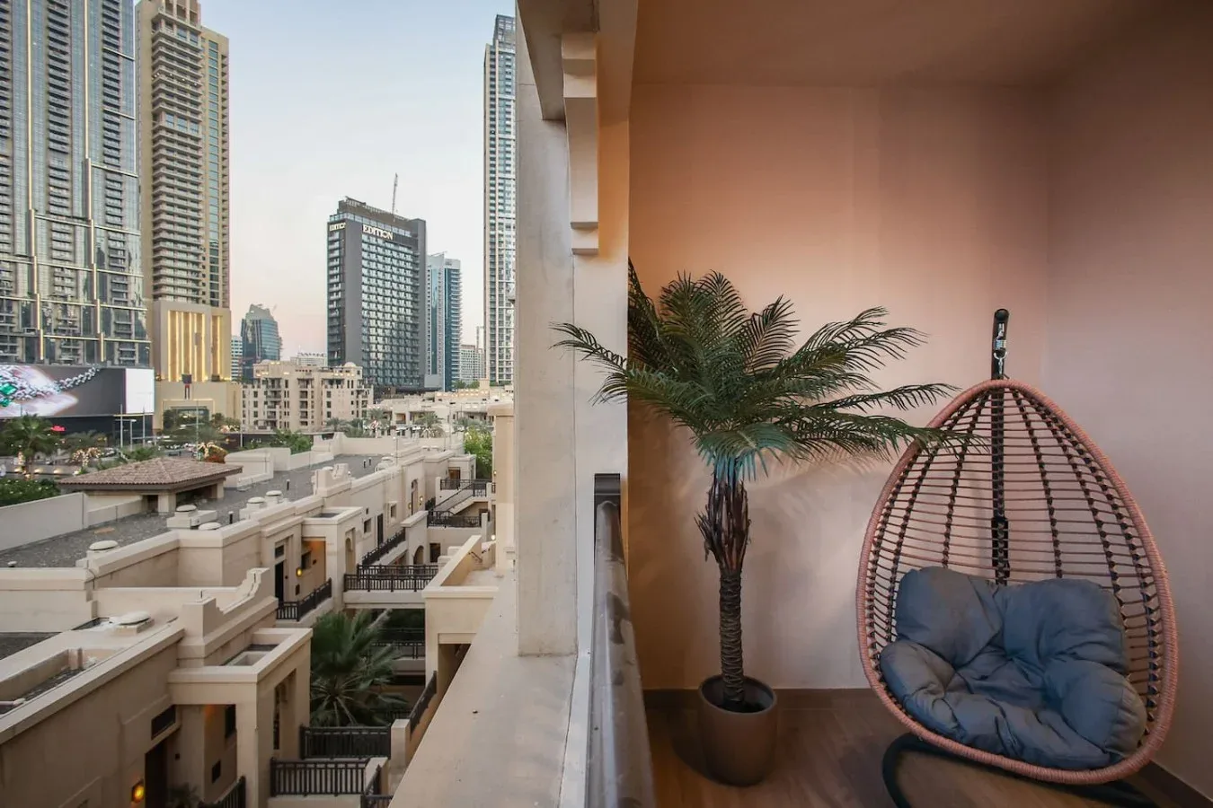 Una zona de estar en un balcón da a edificios cercanos, con una silla colgante y una palmera en maceta.
