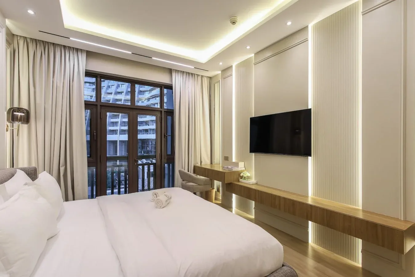 Un dormitorio luminoso cuenta con una cama grande, un televisor en la pared y ventanas altas con cortinas.