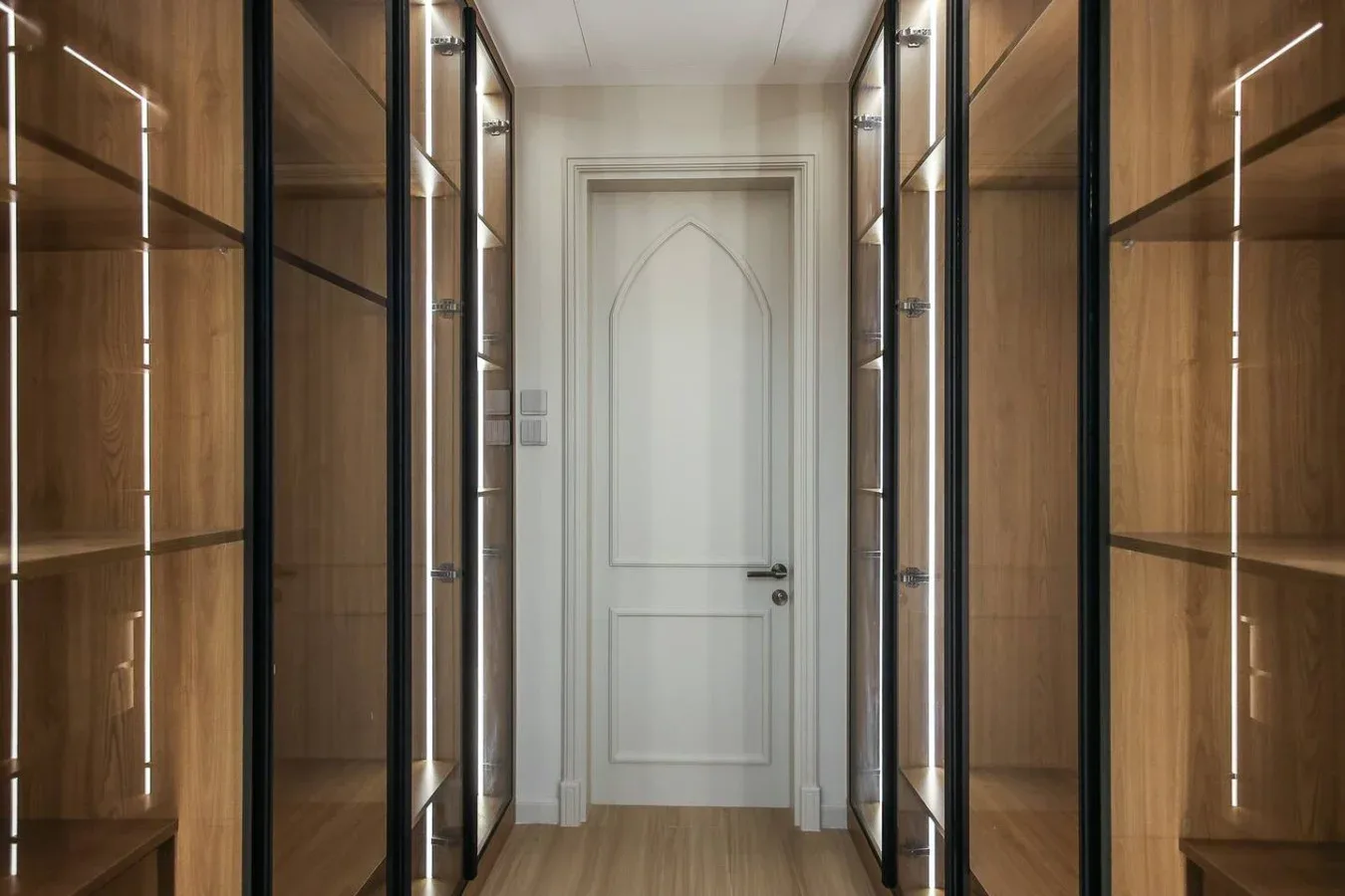 Un pasillo flanqueado por armarios de madera iluminados con puertas de cristal conduce a una puerta blanca.