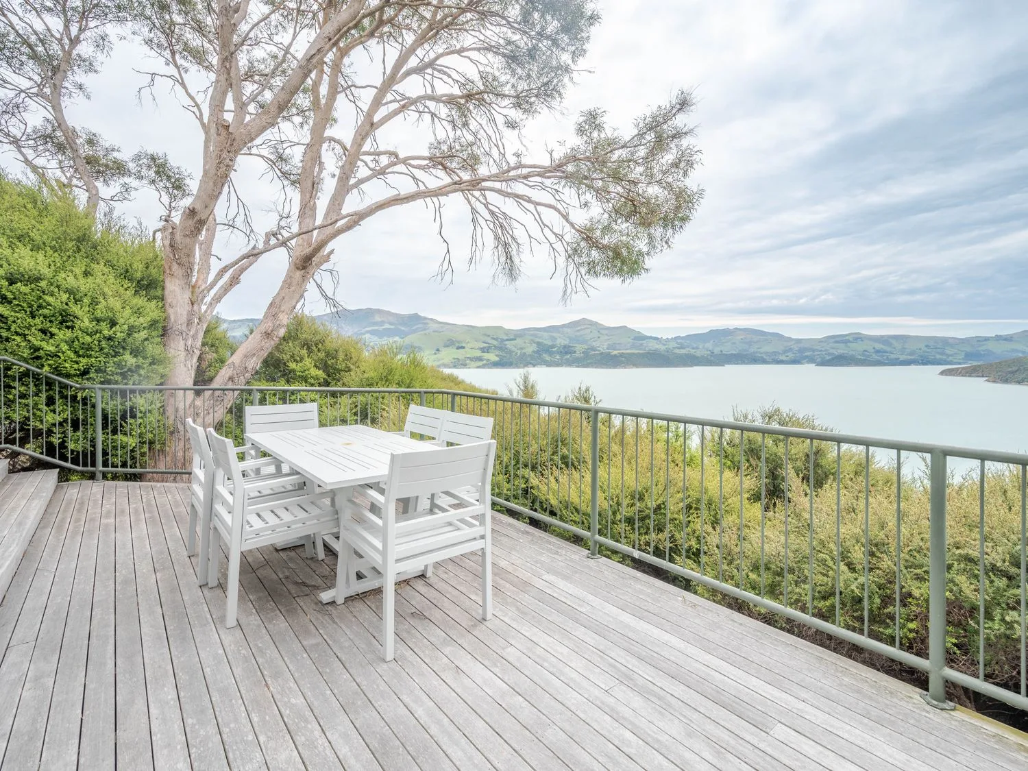 An outdoor deck with a white dining table and six chairs overlooking a body of water and hills at Harbour Haven Akaroa