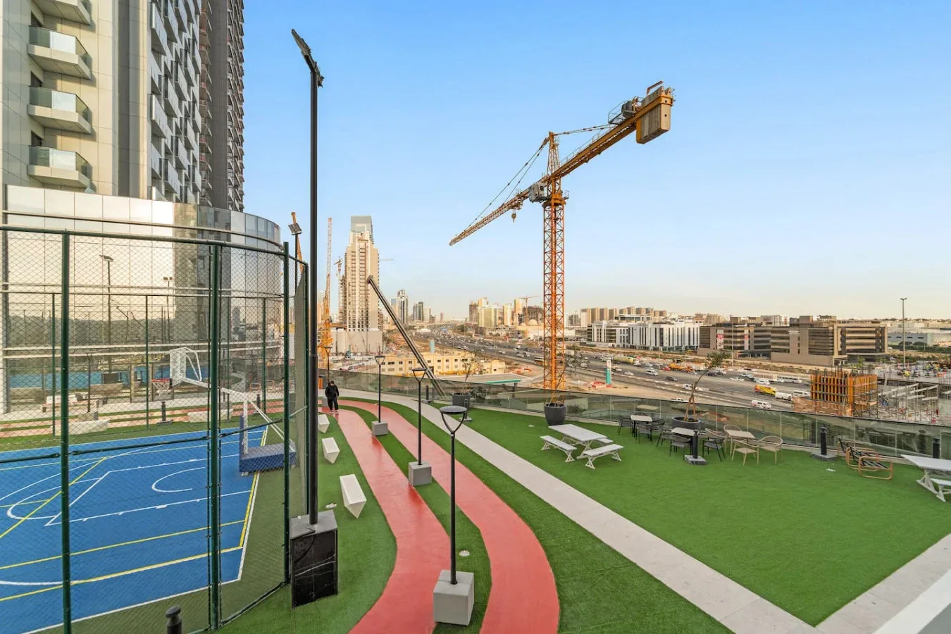Zona recreativa al aire libre con una cancha cercada, pista para correr y grúas de construcción al fondo.