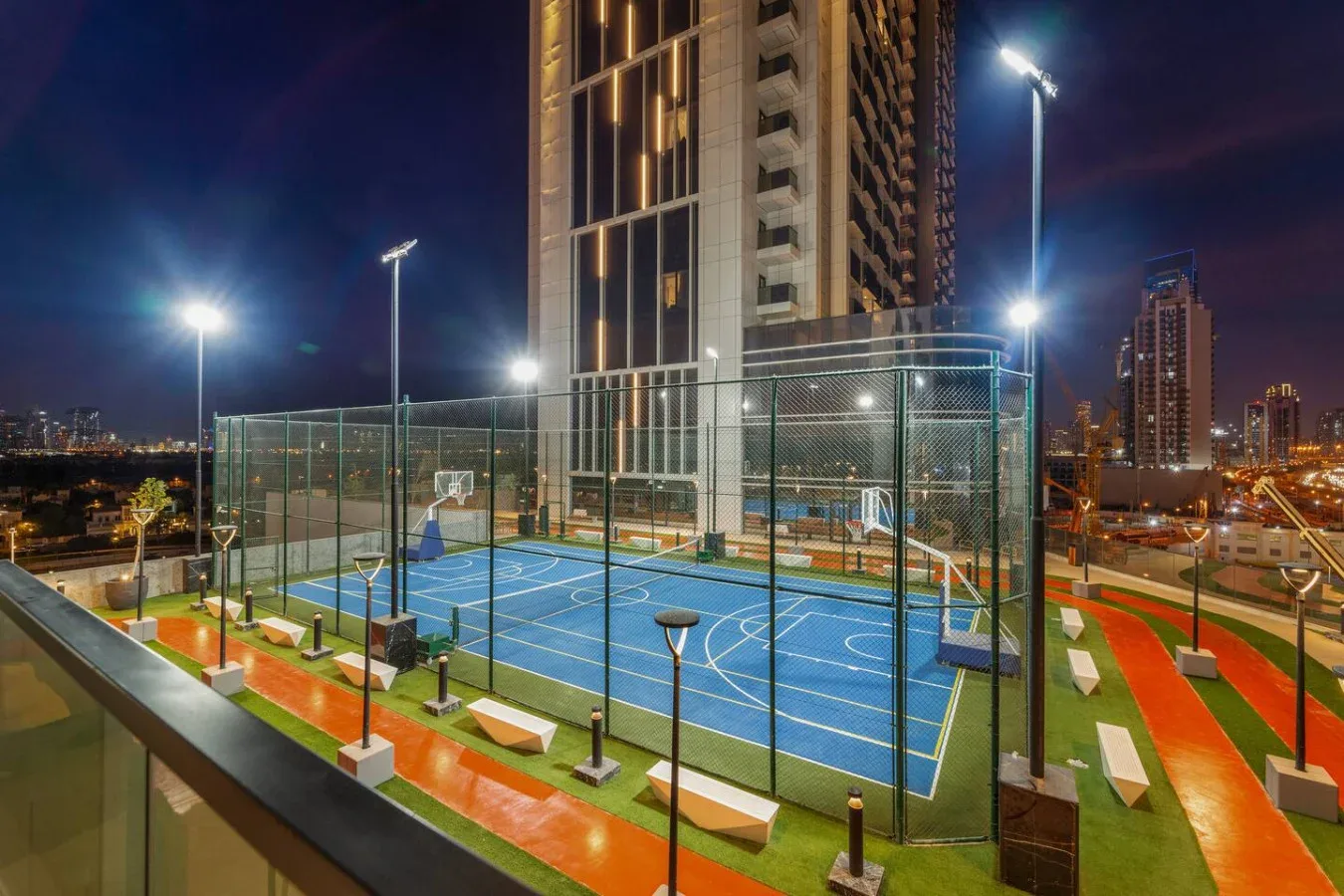 Una cancha deportiva al aire libre con superficies de juego azules y edificios urbanos de noche.