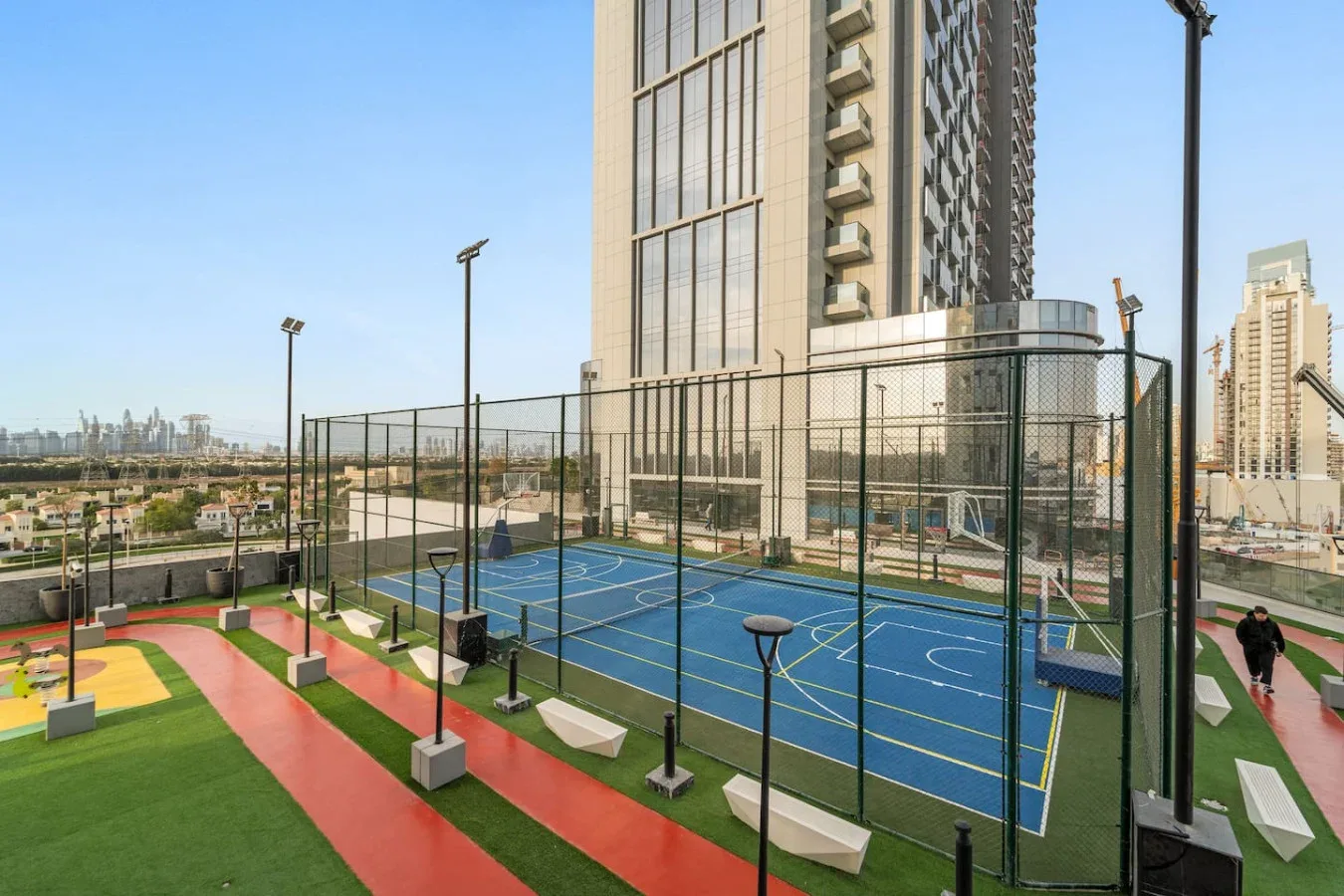 Cancha deportiva al aire libre junto a un edificio alto, con vistas de la ciudad más allá de la cerca.