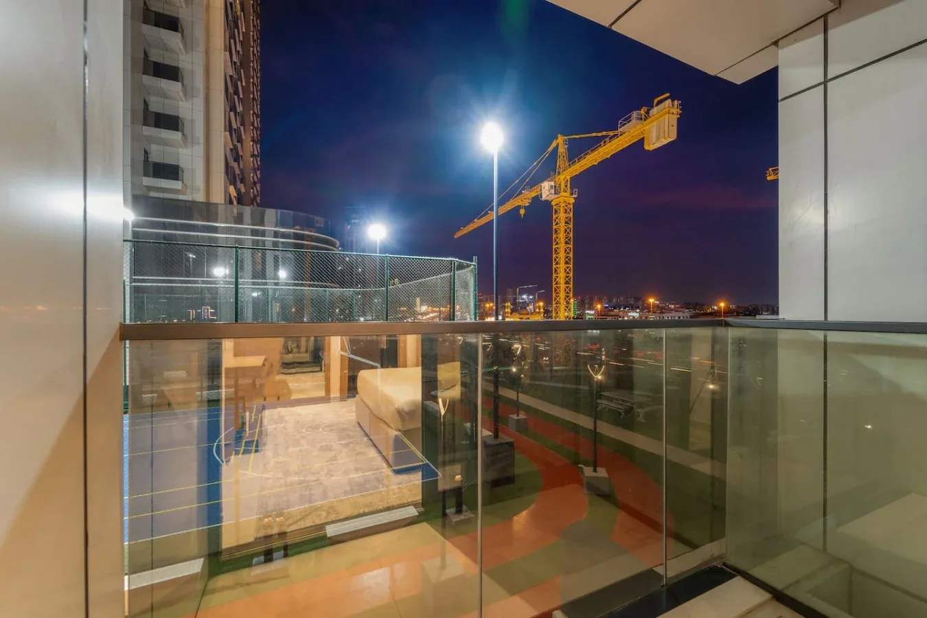 Un balcón de vidrio da a grúas de construcción iluminadas y luces de la ciudad por la noche.