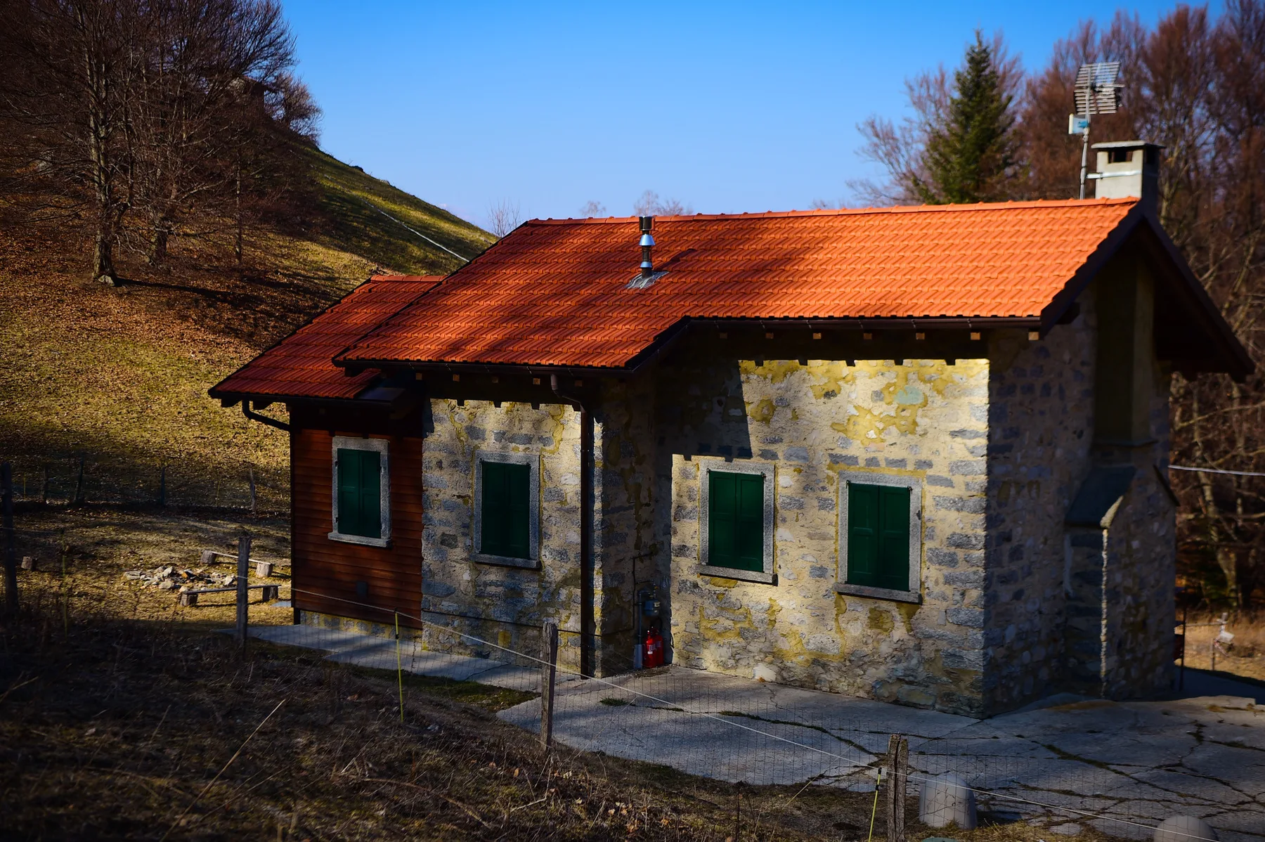 A stone house with a red tiled roof stands beside a wooded hillside.