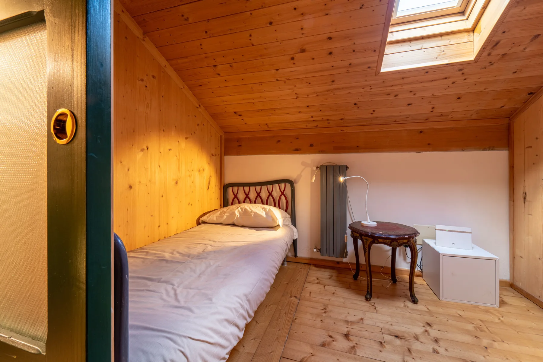 A small bedroom with a bed beneath a sloped wooden ceiling and skylight.