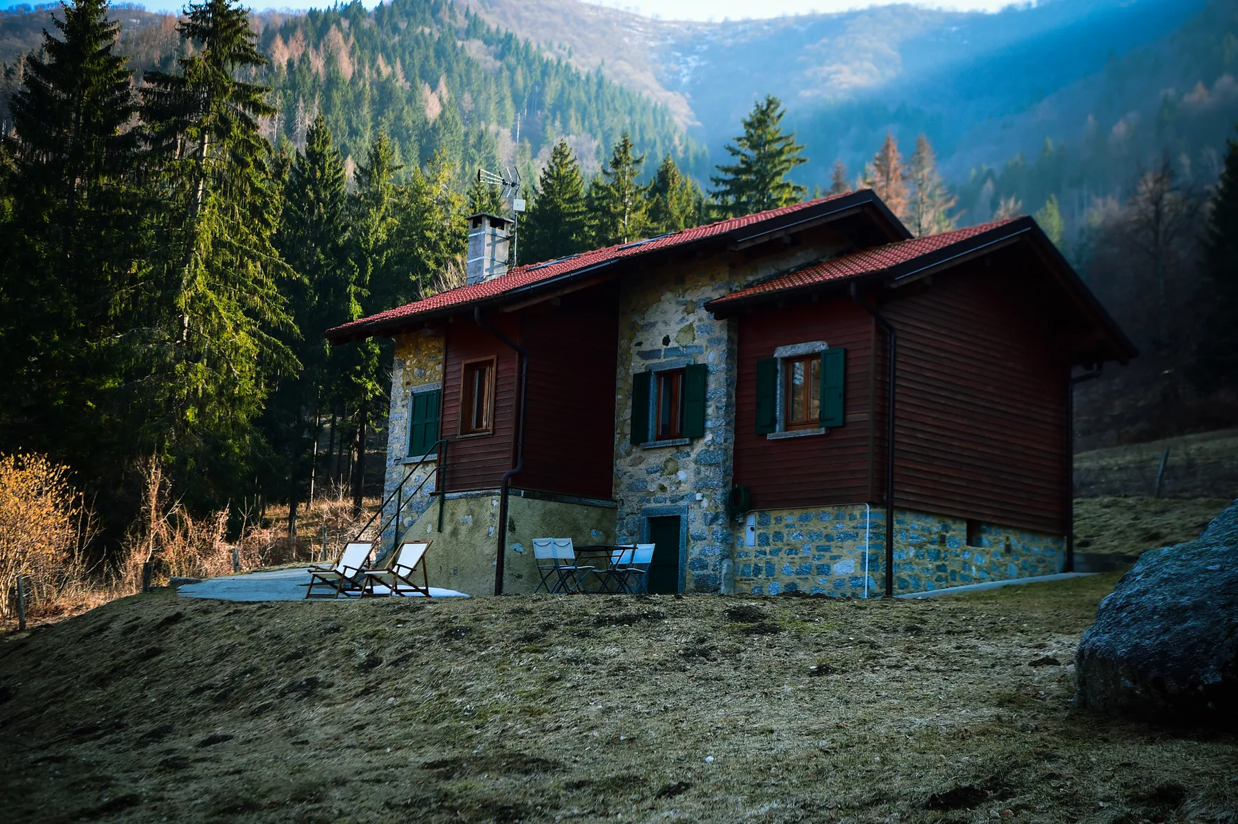 A rustic cabin sits in a mountain clearing, surrounded by evergreen trees.