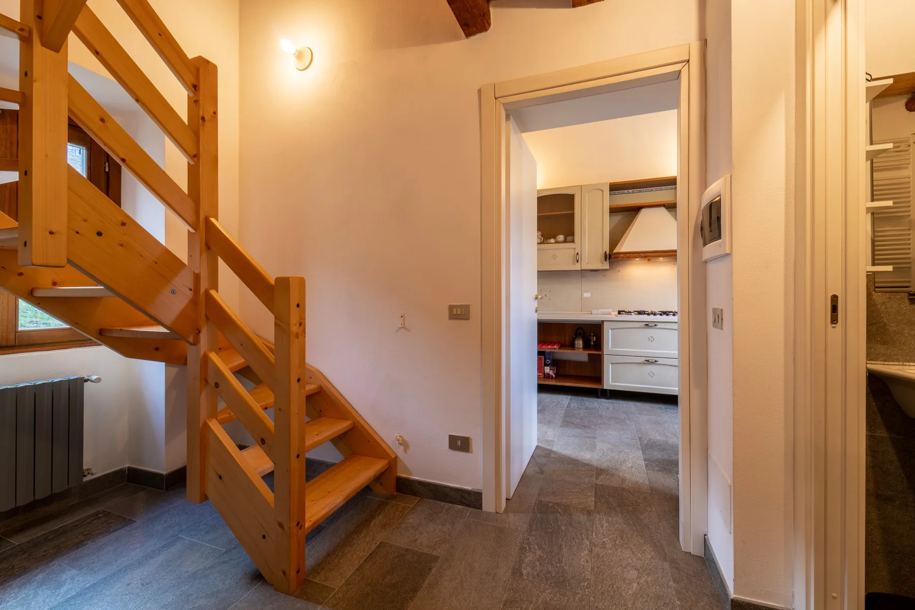 A room with a wooden staircase and a kitchen visible through the doorway.