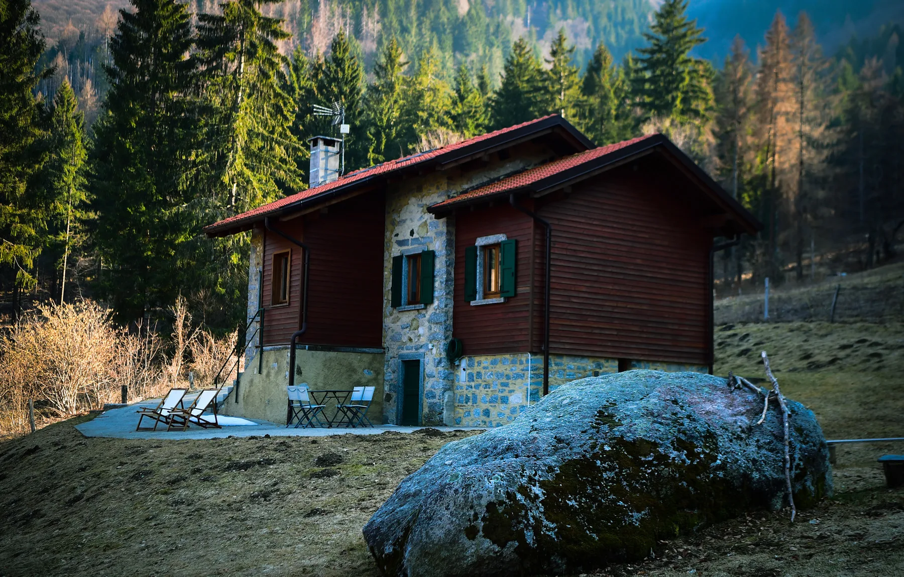 A wooden cabin stands among trees beside a large boulder.