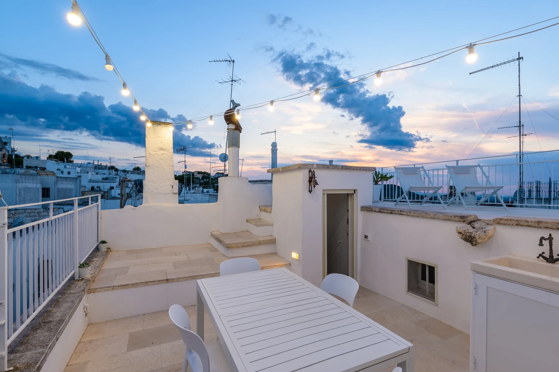 A rooftop terrace with a white dining table and string lights at dusk.