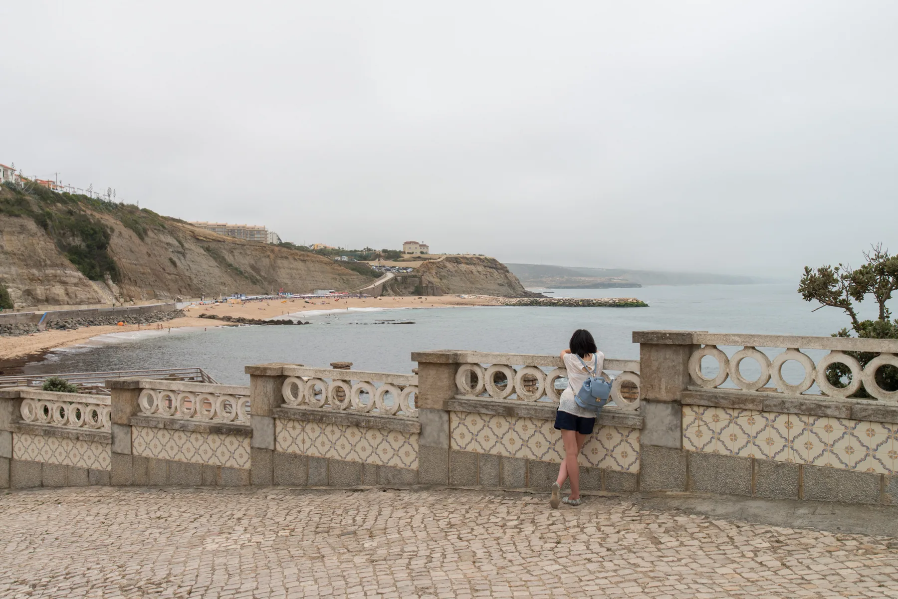Un paseo junto al mar da a una bahía, con una persona apoyada en la barandilla de piedra.
