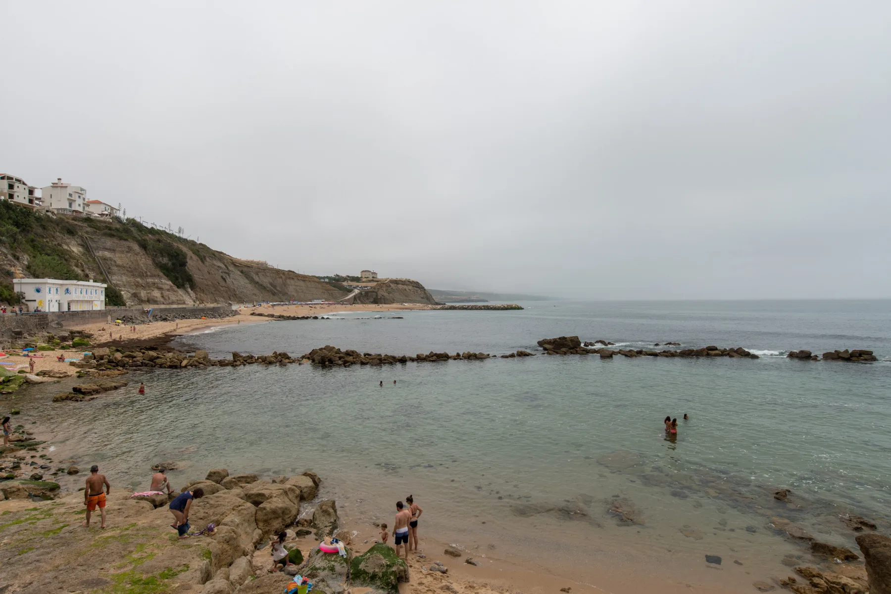 Una playa rocosa con bañistas en el agua y edificios a lo largo de la costa.