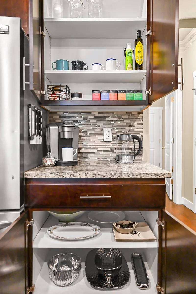 A kitchen counter with a coffee maker beneath open cupboards stocked with mugs and supplies.