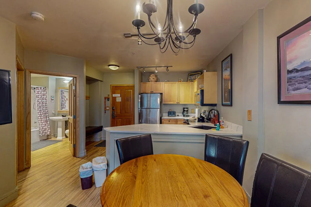 A dining area opens onto a compact kitchen, with a round wooden table in the foreground.