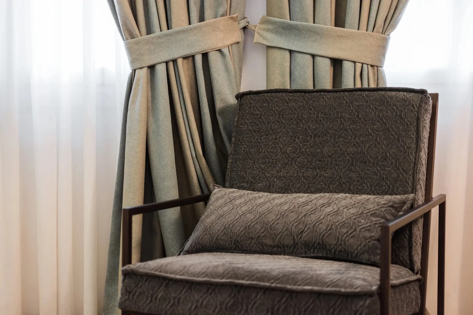 An upholstered armchair sits in front of tied-back curtains.