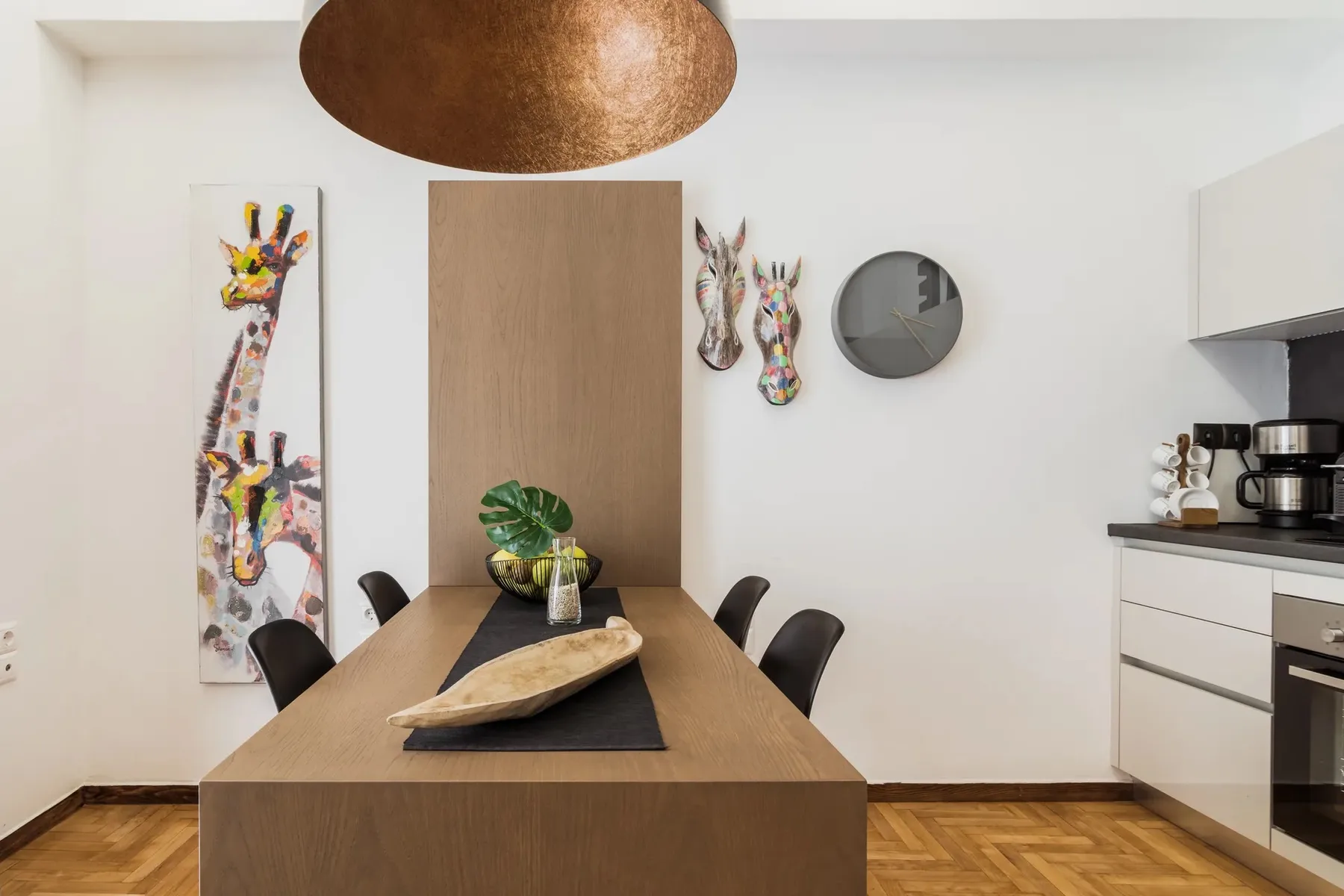 A dining table sits beside a compact kitchen, with colorful wall art behind it.