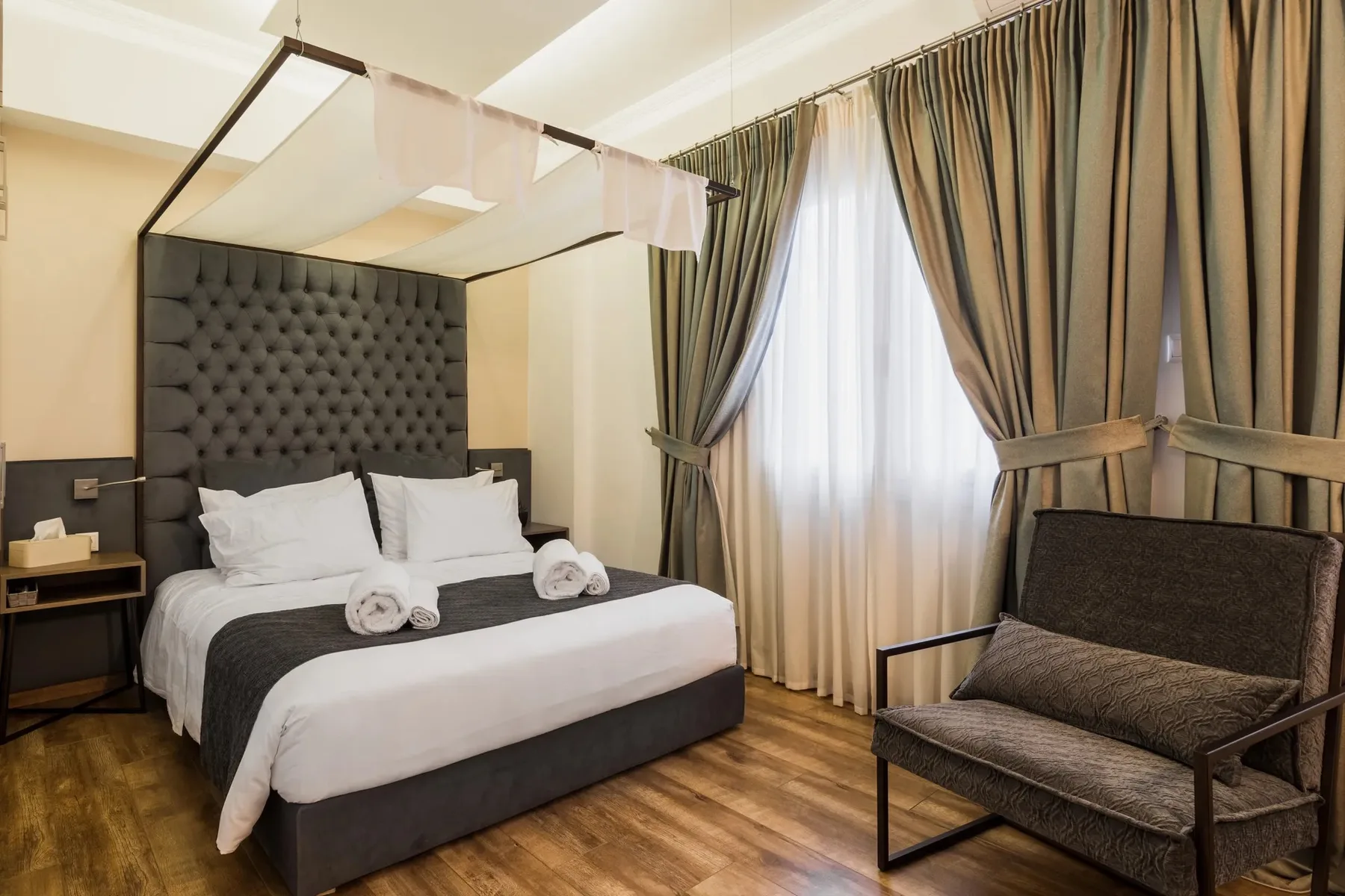A bedroom with a neatly made bed and a chair beside curtained windows.