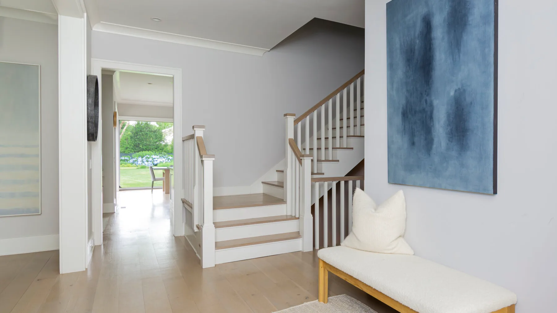 A bright entry hall with a staircase and a cushioned bench beneath blue wall art.