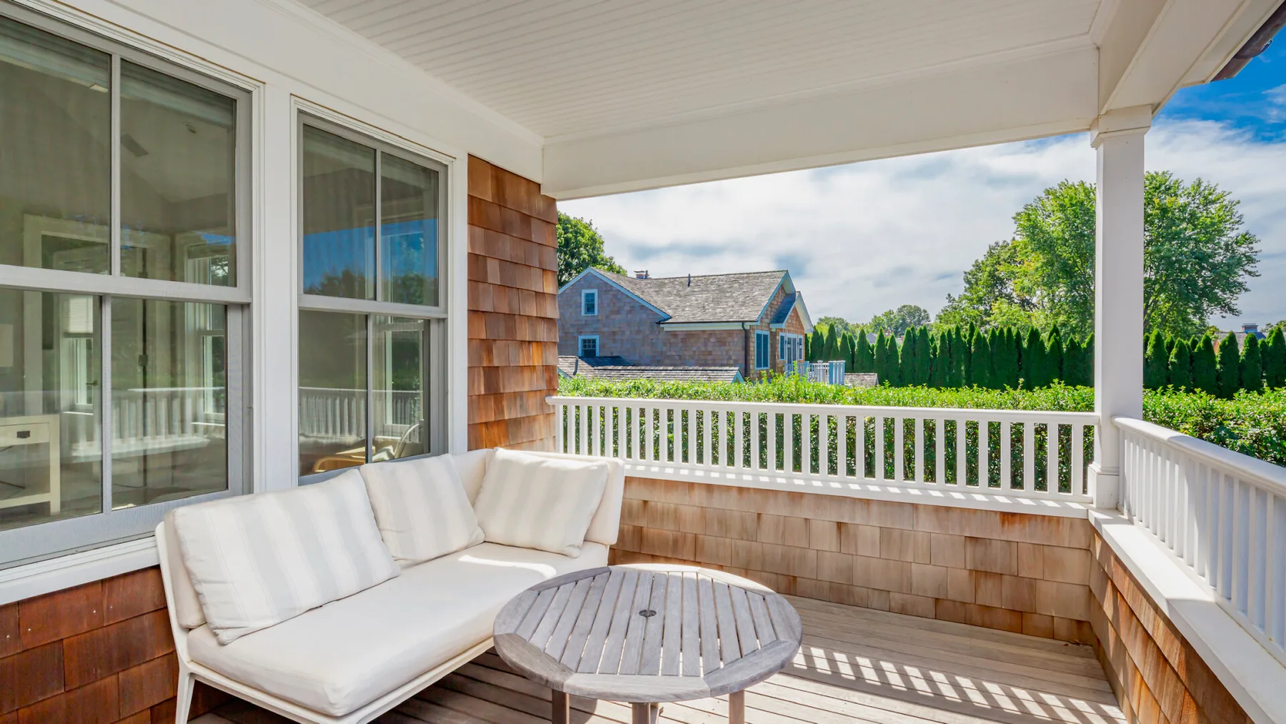 A covered porch with cushioned seating and a round table overlooks a fenced yard.