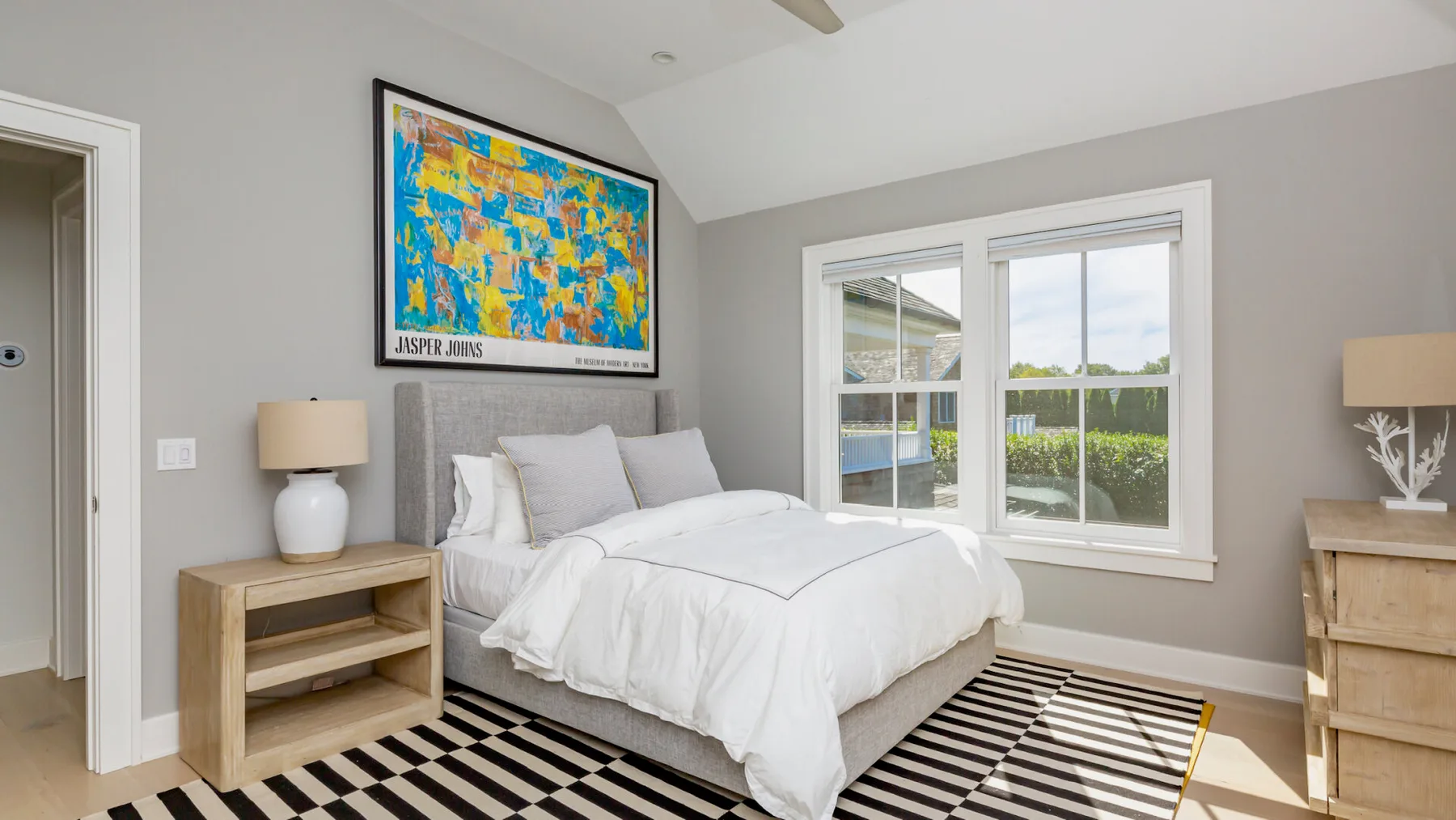 A bedroom with a white bed, colorful wall art, and large windows.
