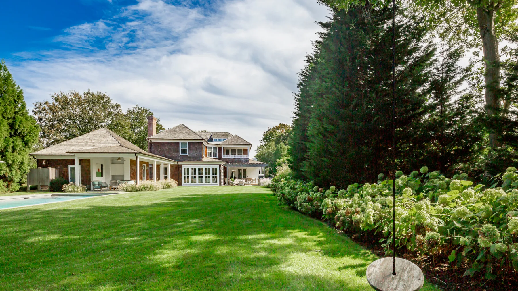 A large house and covered patio overlook a green lawn and garden.