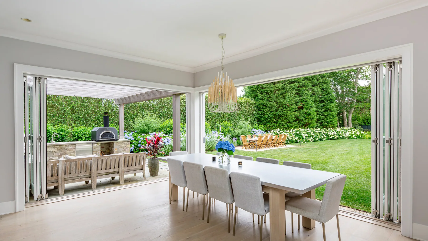 A dining area opens onto a garden, with a long table and surrounding seating.
