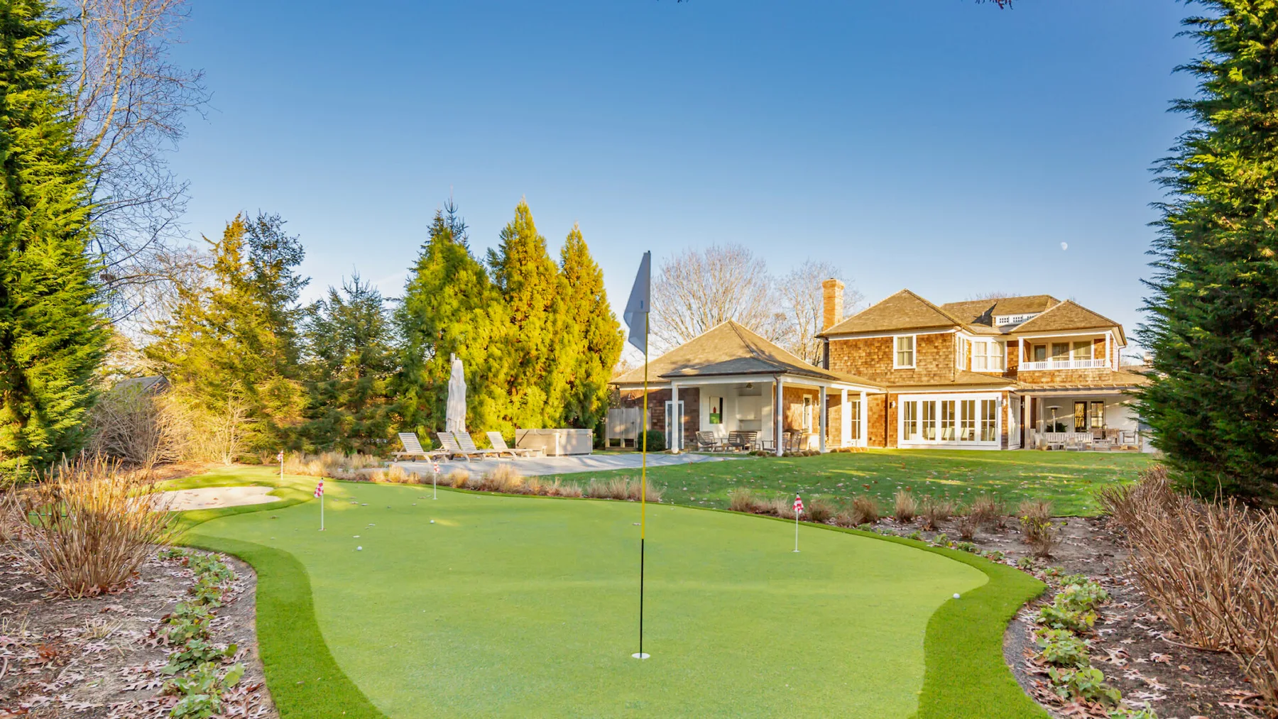 A landscaped golf green with a flag stands in front of a large house.
