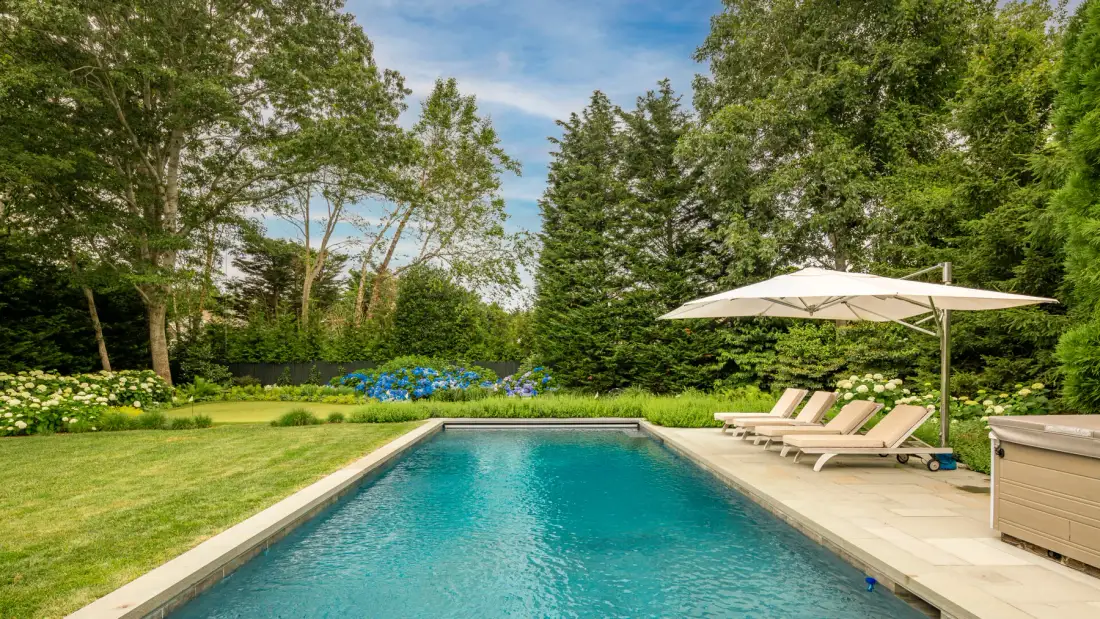 An outdoor swimming pool sits beside a lawn, with lounge seating and an umbrella.