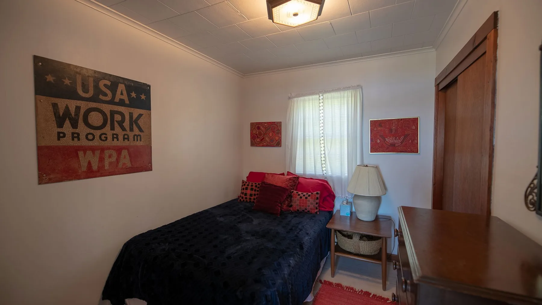 Un dormitorio con una cama, una mesita de noche y una ventana con cortinas transparentes.