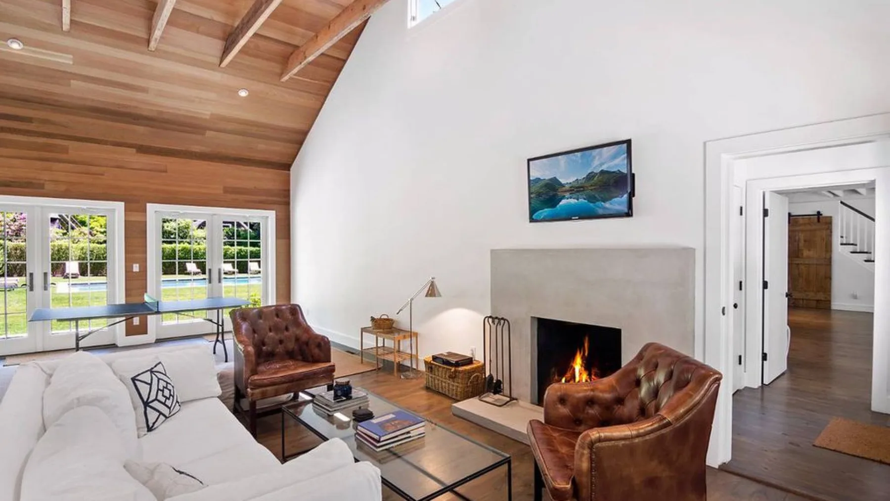 A bright living room with a fireplace, seating, and large windows beneath a sloped wood ceiling.