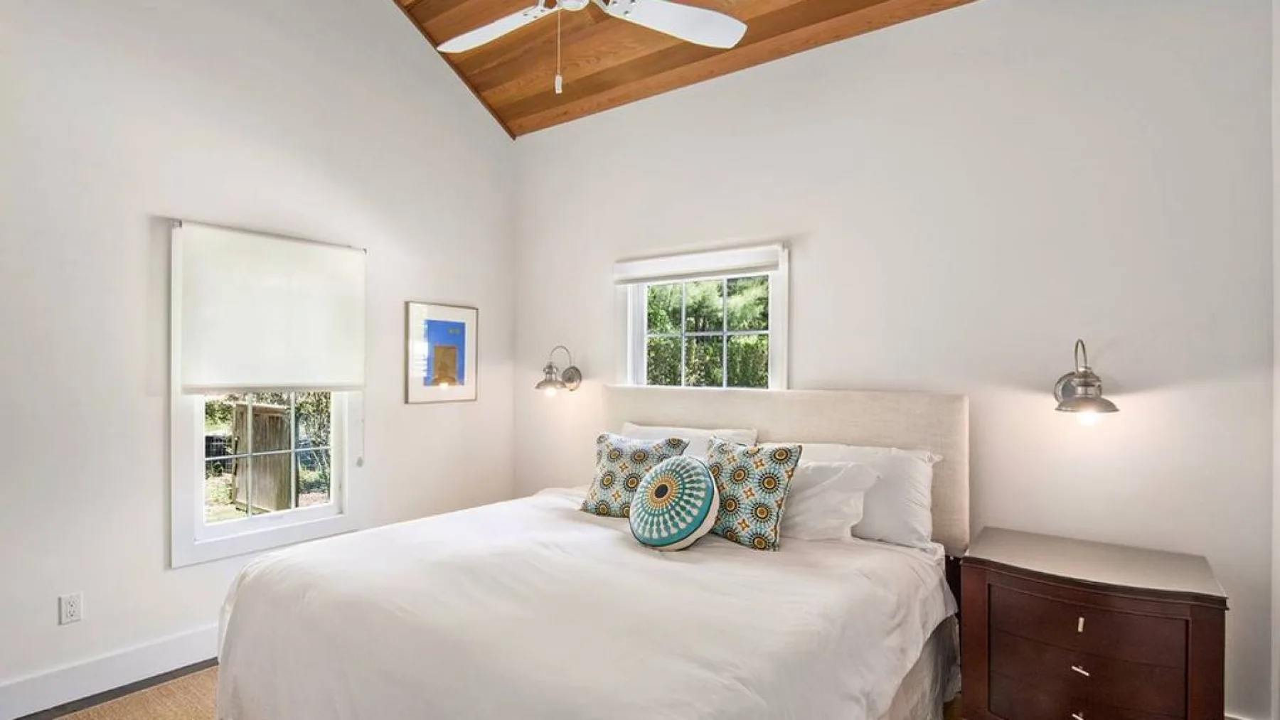A bright bedroom with a bed, patterned pillows, and windows.