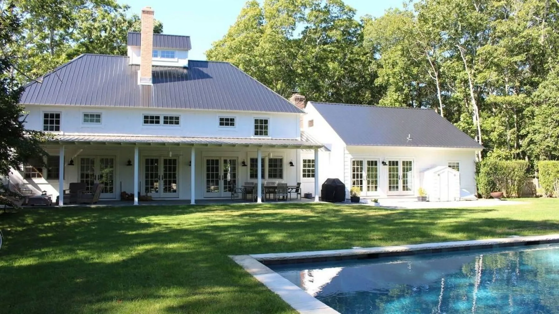 A large white house stands beside a swimming pool on a green lawn.