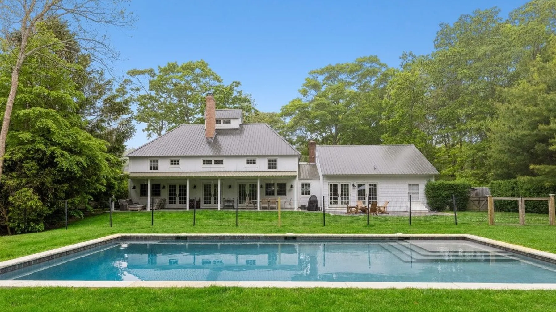 Una casa blanca se alza detrás de una piscina, rodeada de árboles.