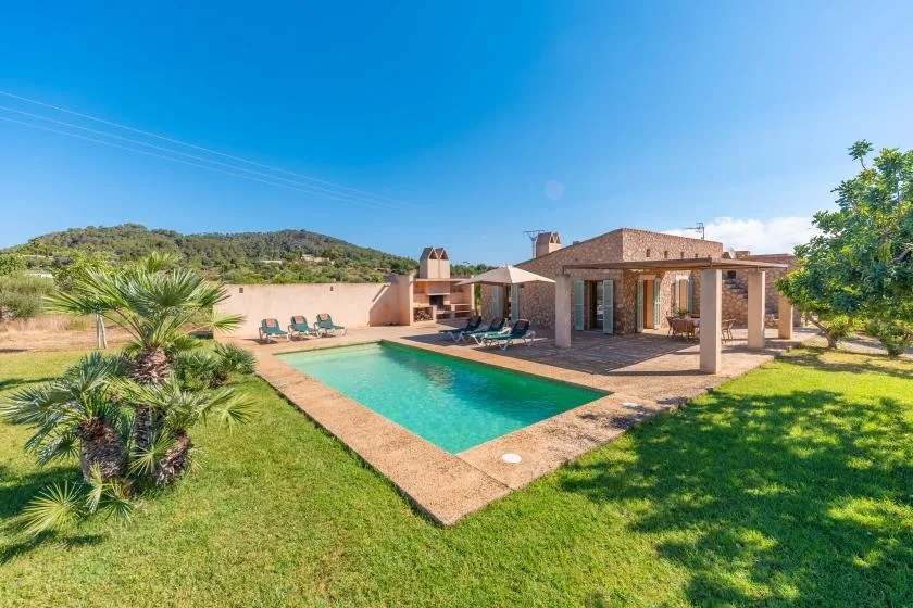 A swimming pool and stone house sit amid a sunny landscaped yard.