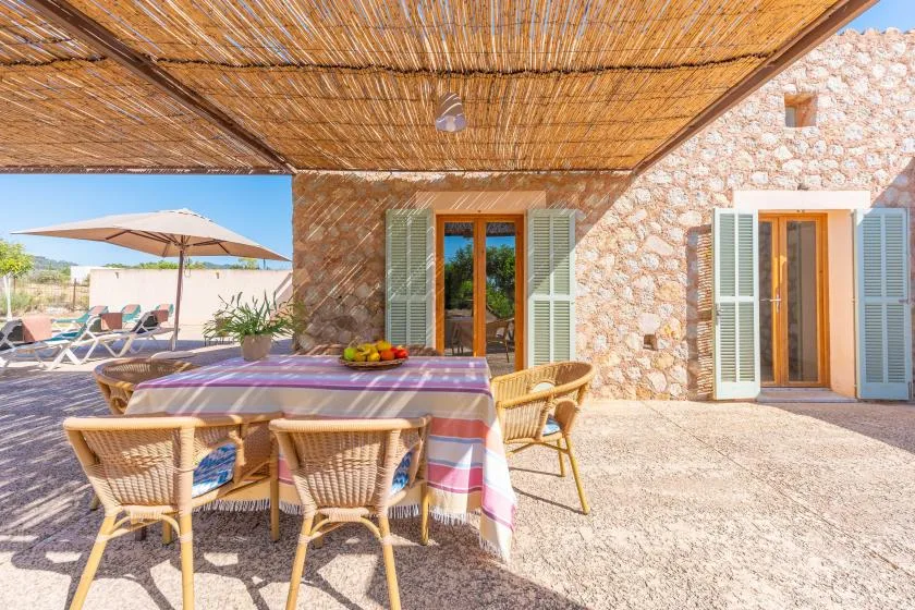 A shaded outdoor dining area with a table and wicker seating beside a stone building.