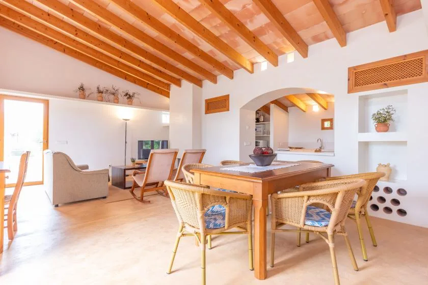 A bright dining and living area with a wooden table and woven seating beneath exposed ceiling beams.