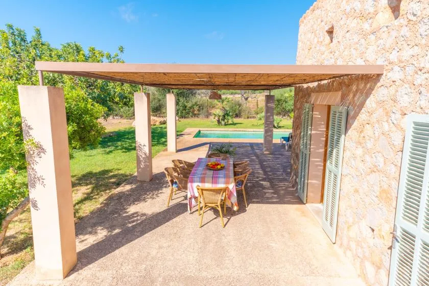 A shaded outdoor dining area beside a stone building overlooks a garden and swimming pool.