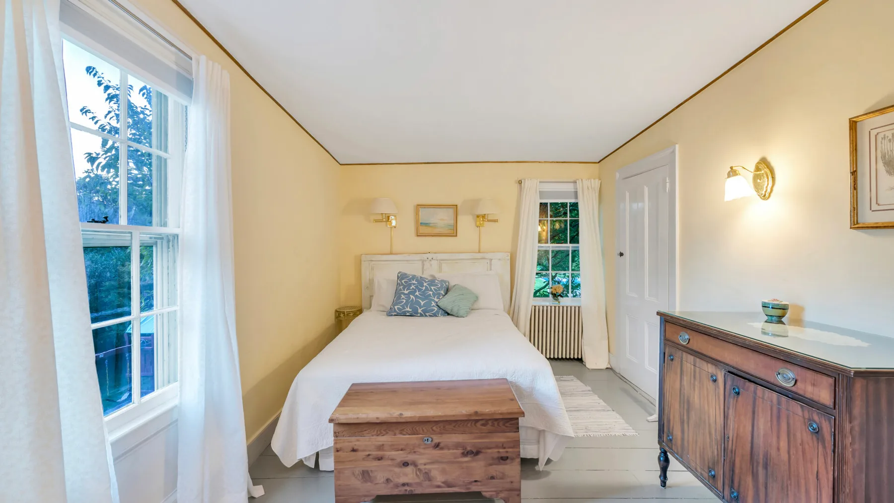 Un dormitorio luminoso con una cama, un baúl de madera y ventanas grandes.