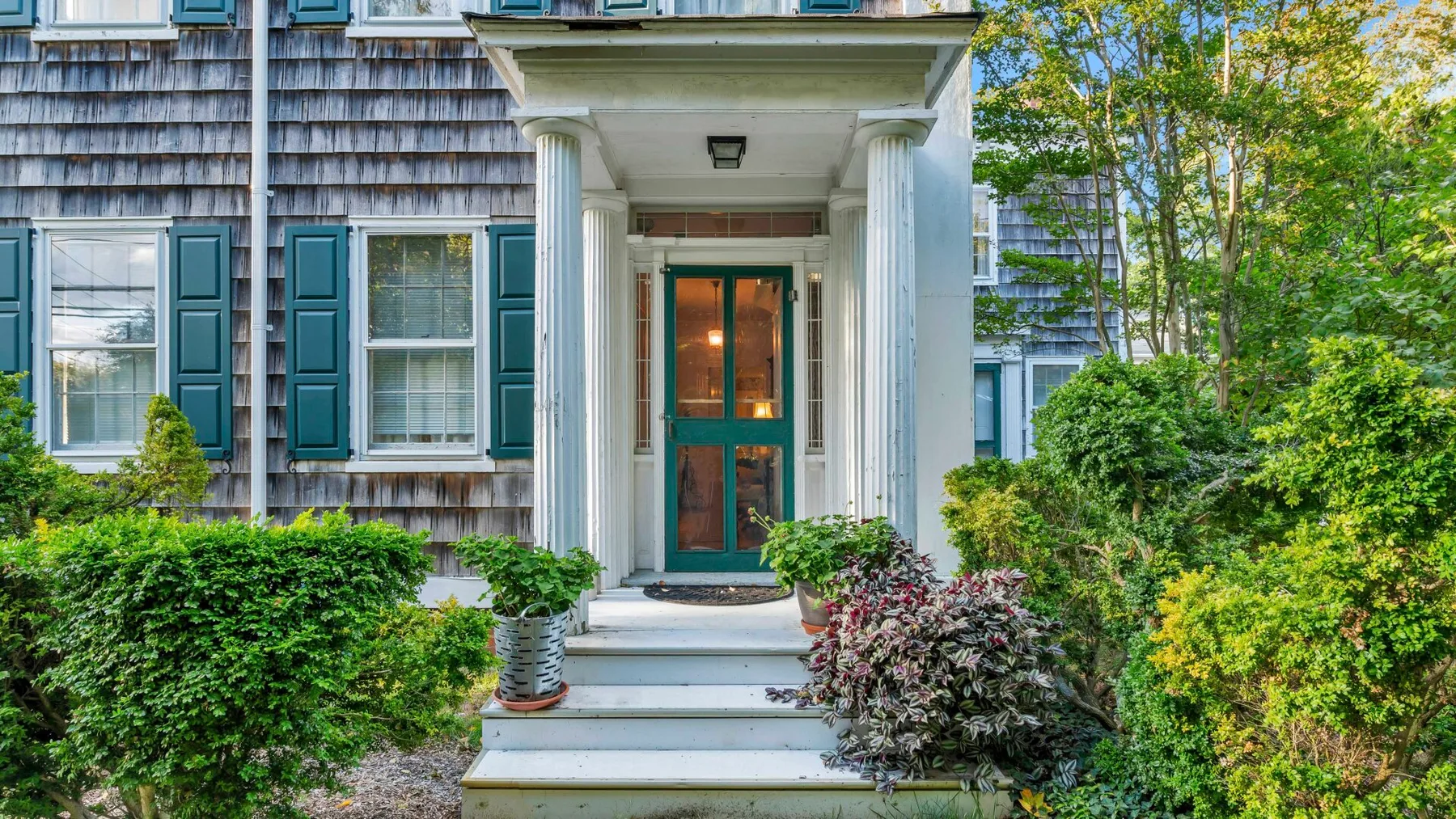 Entrada de una casa con puerta acristalada, columnas blancas y arbustos alrededor.