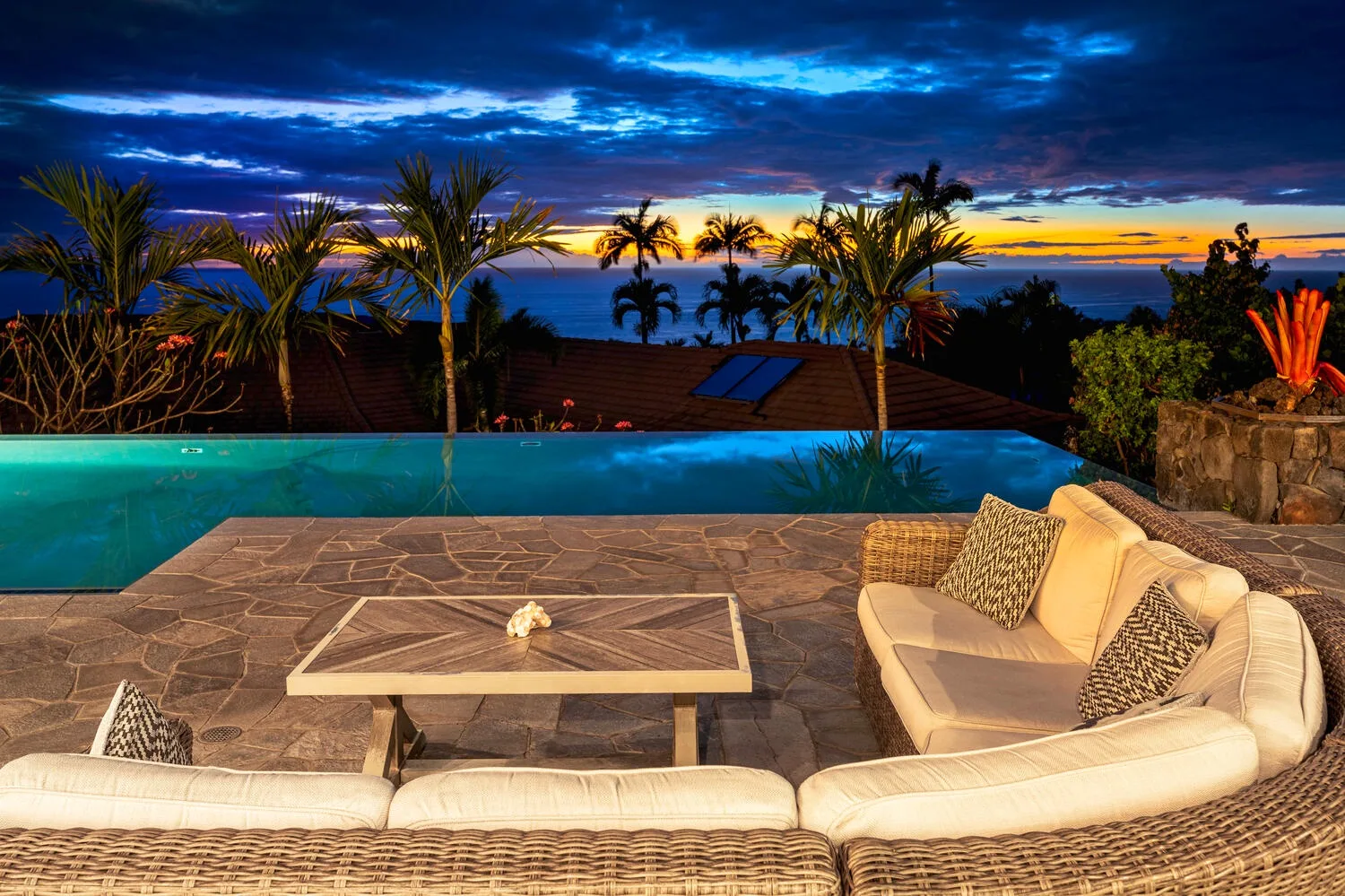 A furnished patio beside a swimming pool overlooks palm trees and a colorful sunset.