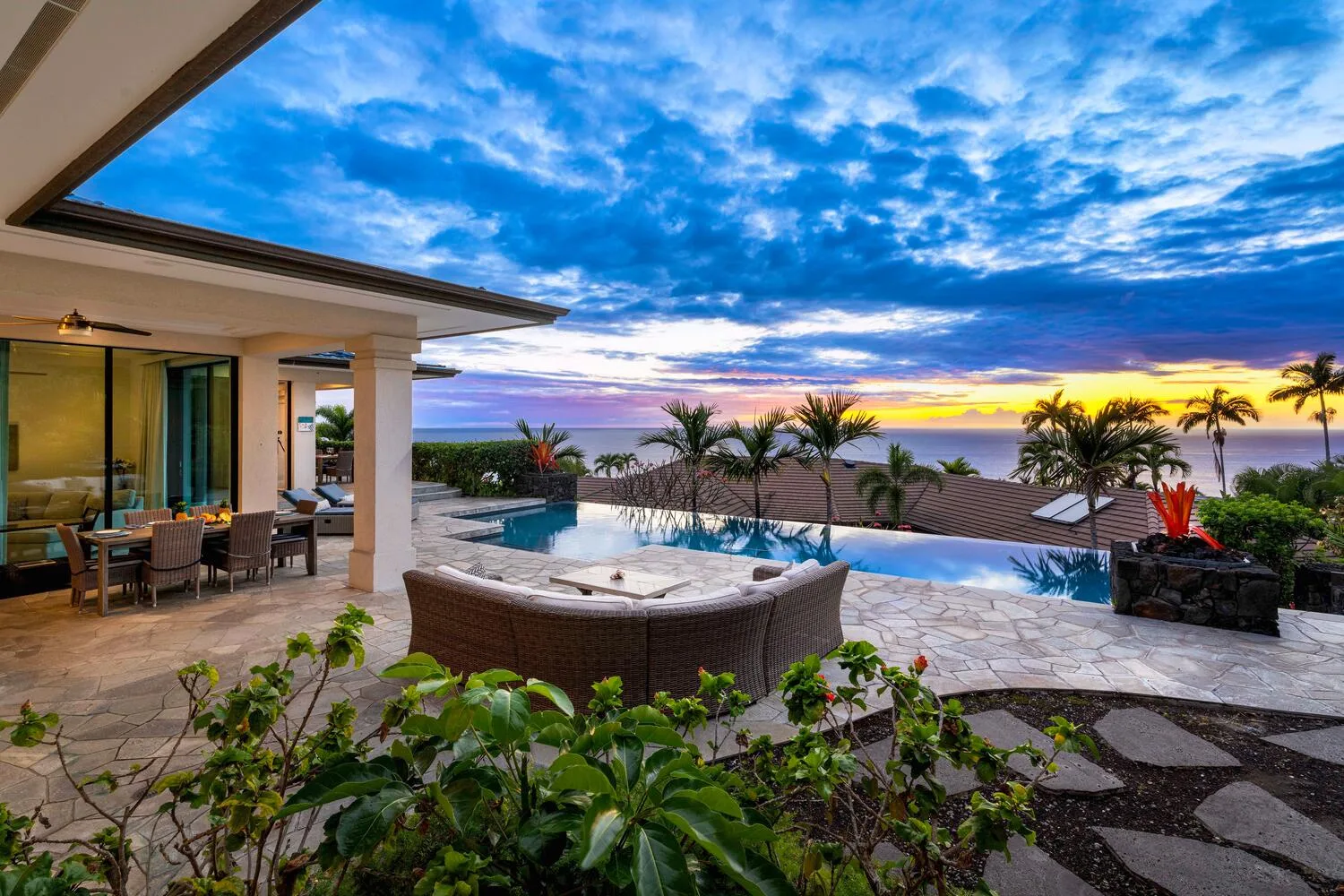 An outdoor patio with a pool overlooks the ocean beneath a colorful sunset sky.
