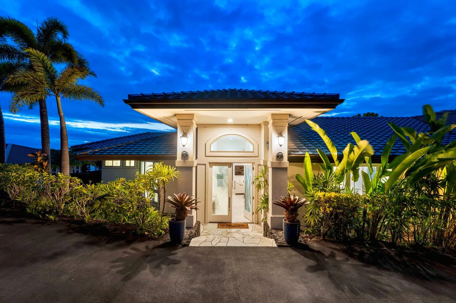A brightly lit house entrance is framed by tropical plants beneath a vivid blue evening sky.