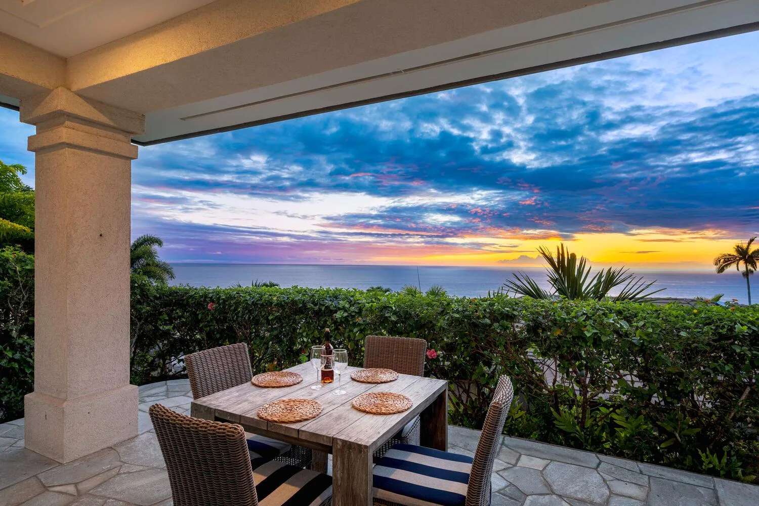 A covered patio dining area overlooks greenery and a colorful sunset over the water.