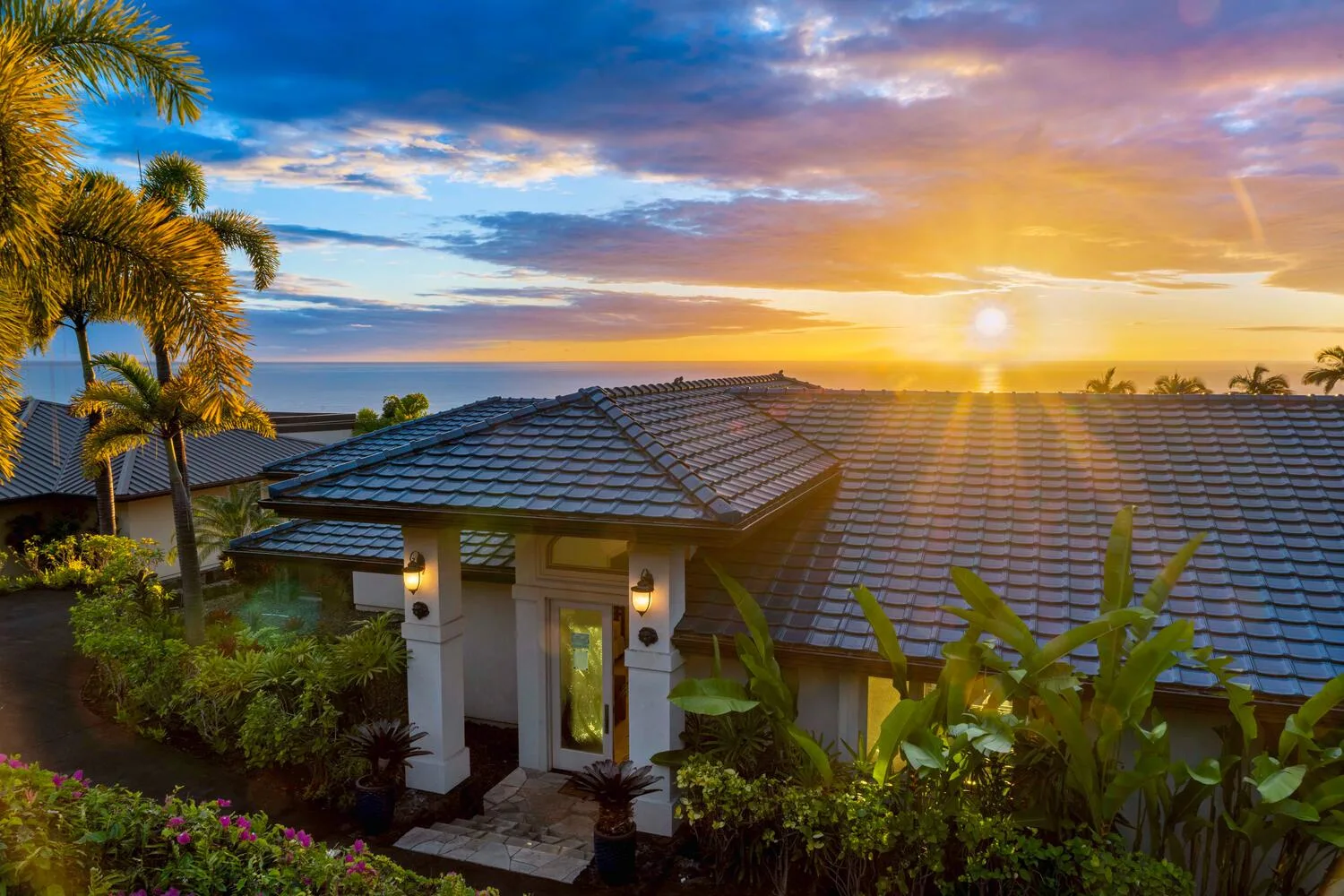A tiled-roof house sits among tropical plants beneath a colorful sunset sky.